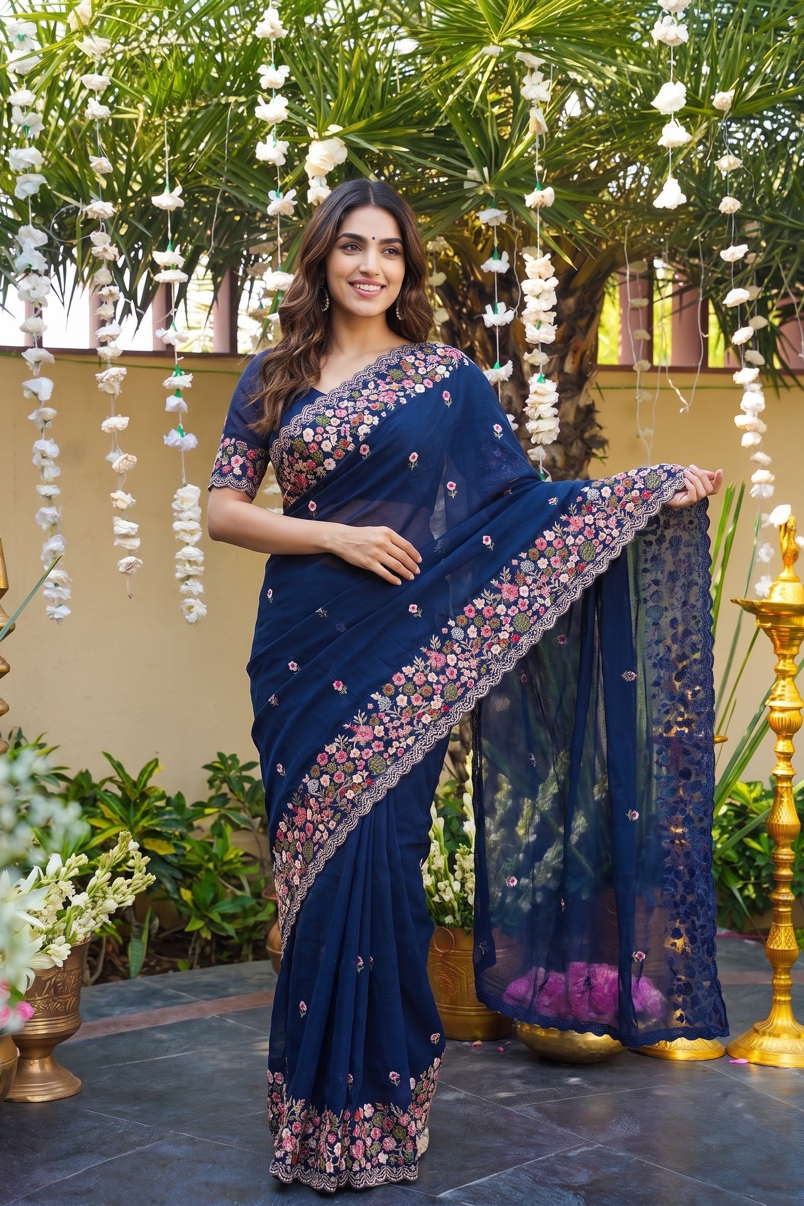 A woman models a navy blue saree with intricate floral embroidery along the border and pallu, set against a decorated outdoor backdrop with hanging white flowers and brass lamps.