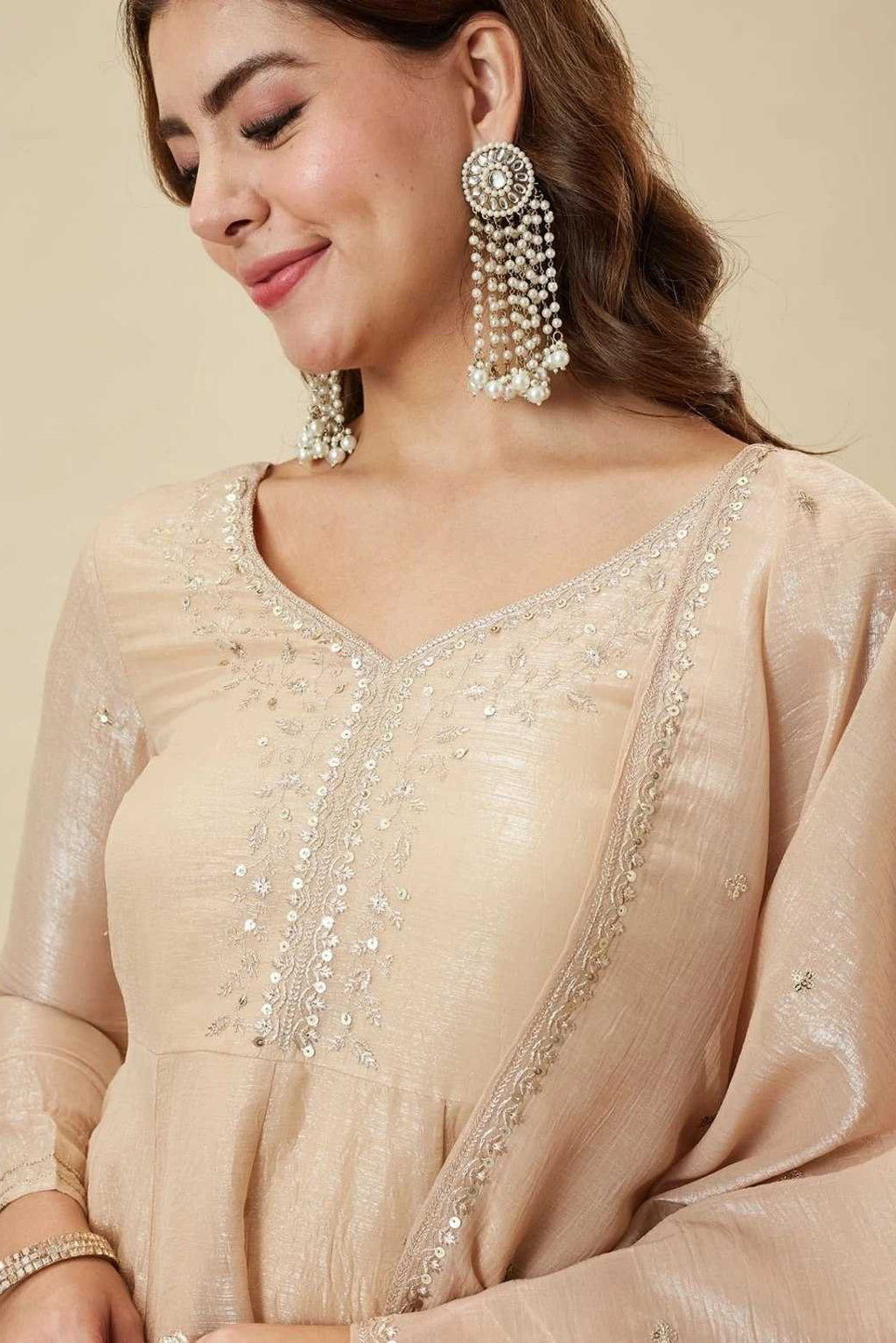 Woman wearing a beige embroidered outfit with matching earrings on a beige background