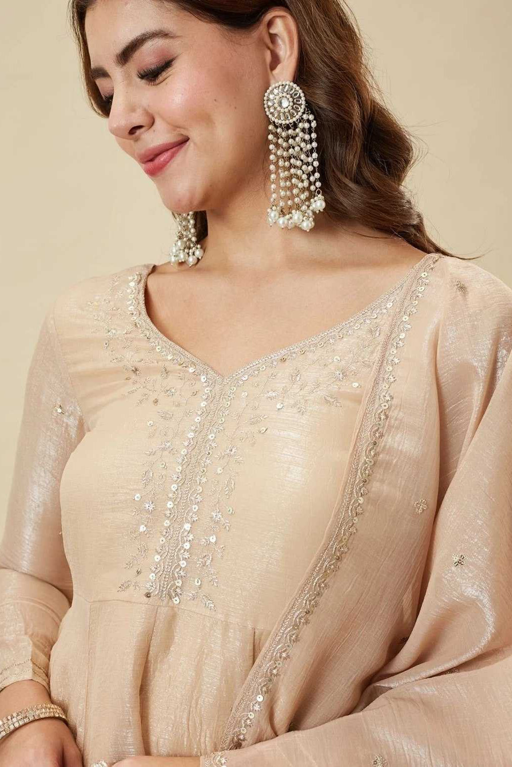 Woman wearing a beige embroidered outfit with matching earrings on a beige background