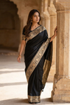 Woman in a black saree with gold patterns standing against a stone architectural background