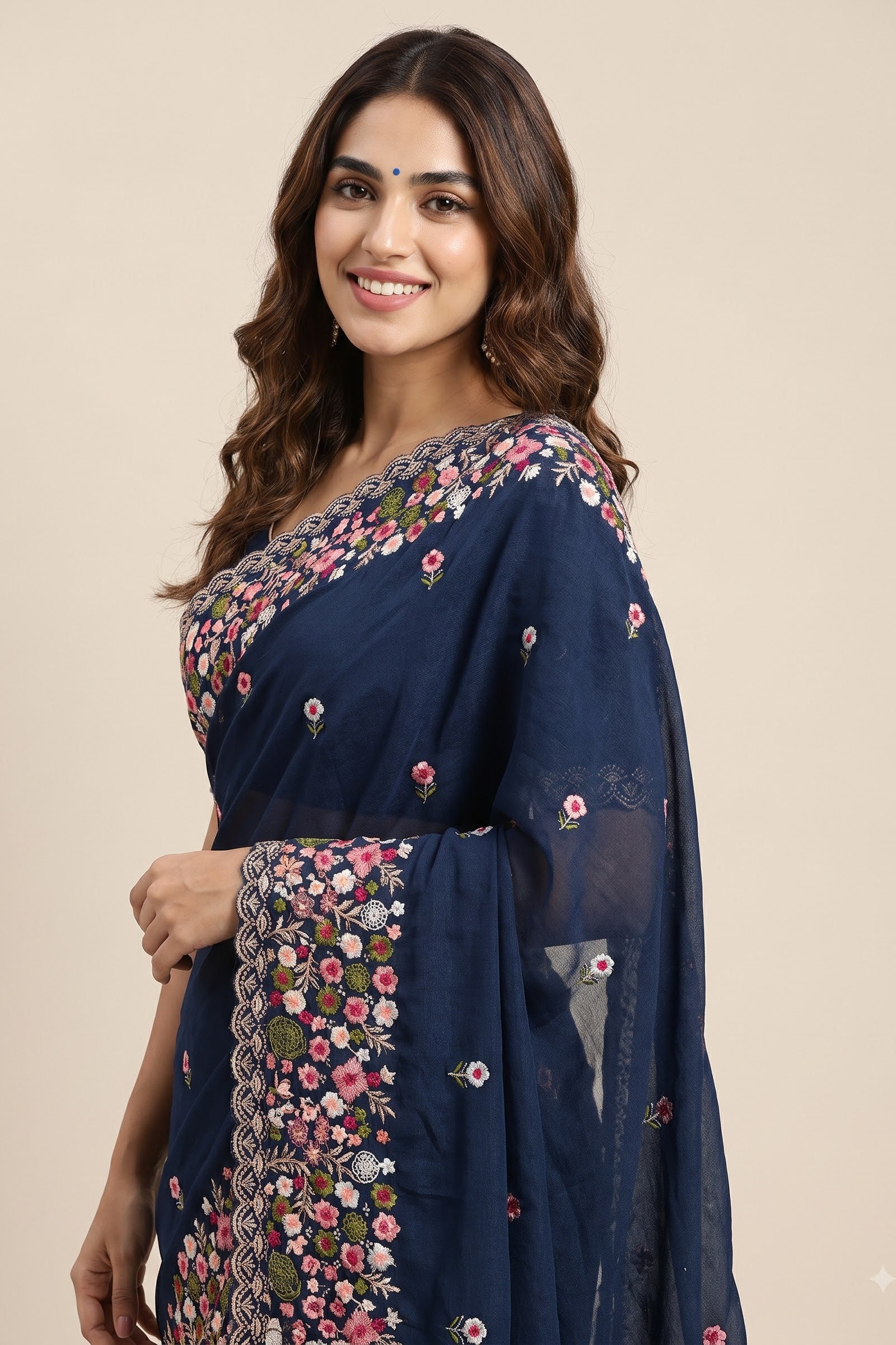 A woman modeling a navy blue saree with intricate floral embroidery and border detailing, set against a solid beige background.