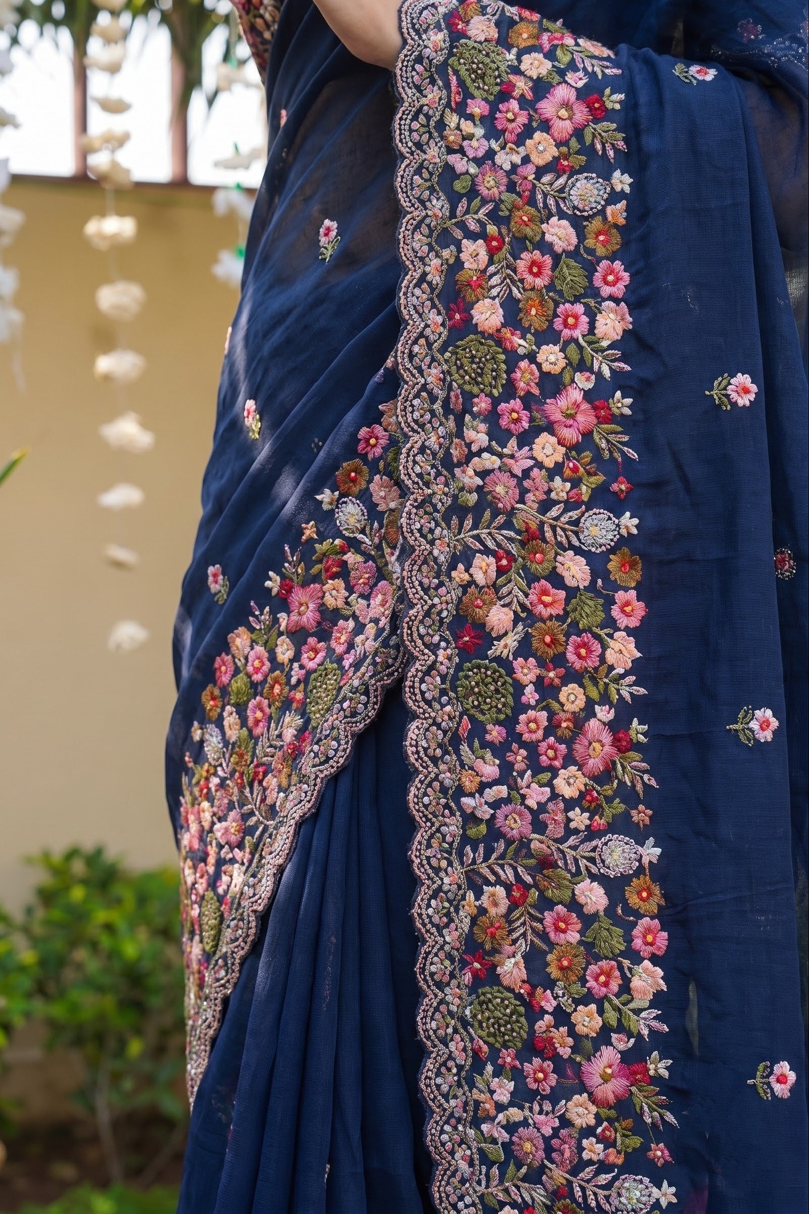A close-up of a navy blue saree with intricate multicolor floral embroidery and a scalloped border, shown on a model in an outdoor garden setting.