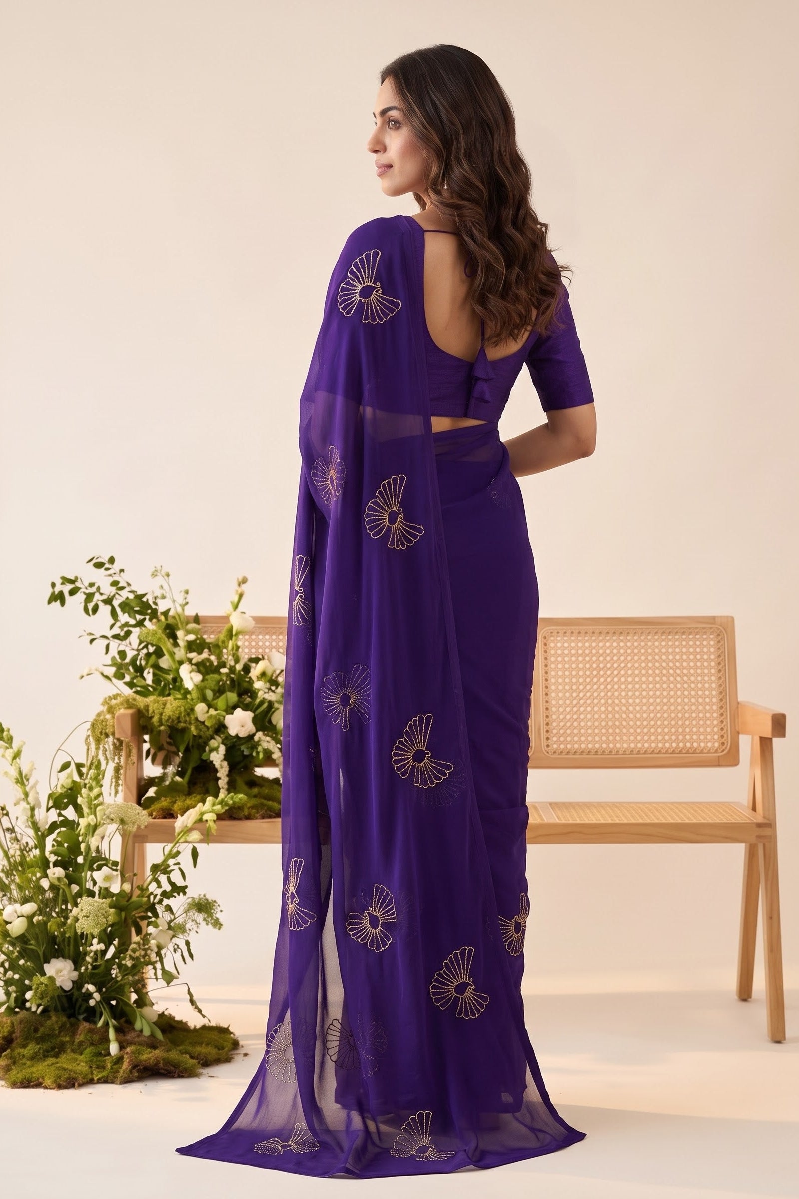 A woman stands with her back to the camera wearing a deep purple saree with gold floral embroidery, paired with a matching purple blouse. She is posed in front of a light beige wall with a wooden cane-back chair and decorative greenery.