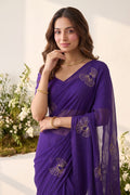A woman wearing a purple georgette saree with gold floral embroidery, standing in front of a white wall with decorative white flowers.
