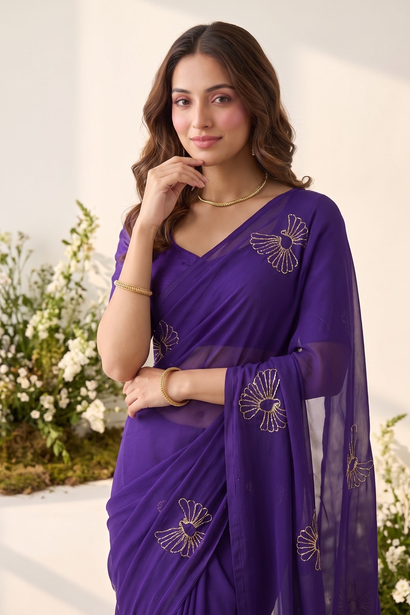 A woman wearing a purple georgette saree with gold floral embroidery, standing in front of a white wall with decorative white flowers.