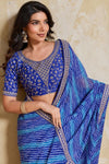 Woman wearing a blue saree with intricate patterns against a beige wall.