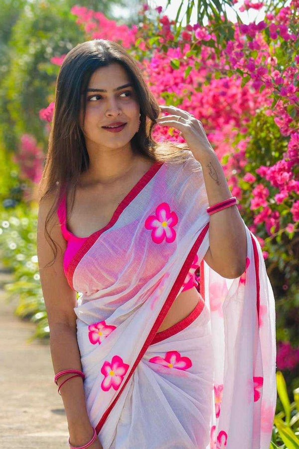 Woman in a white saree with pink floral patterns standing in a garden with pink flowers.