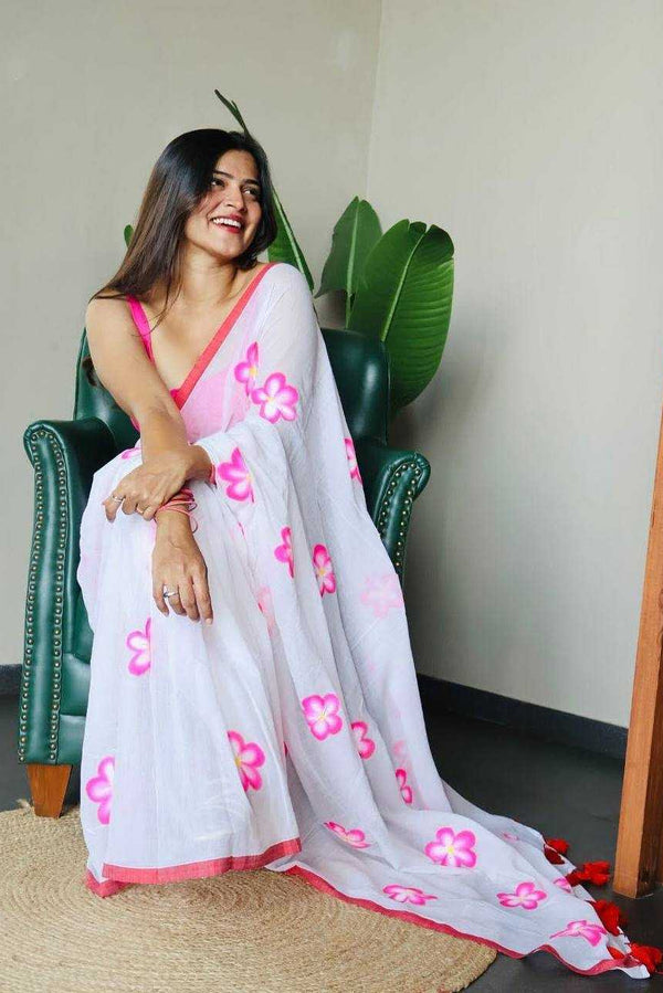 Woman in a white saree with pink floral patterns sitting on a green chair.