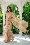 Woman in a beige saree with floral patterns standing in a garden setting.